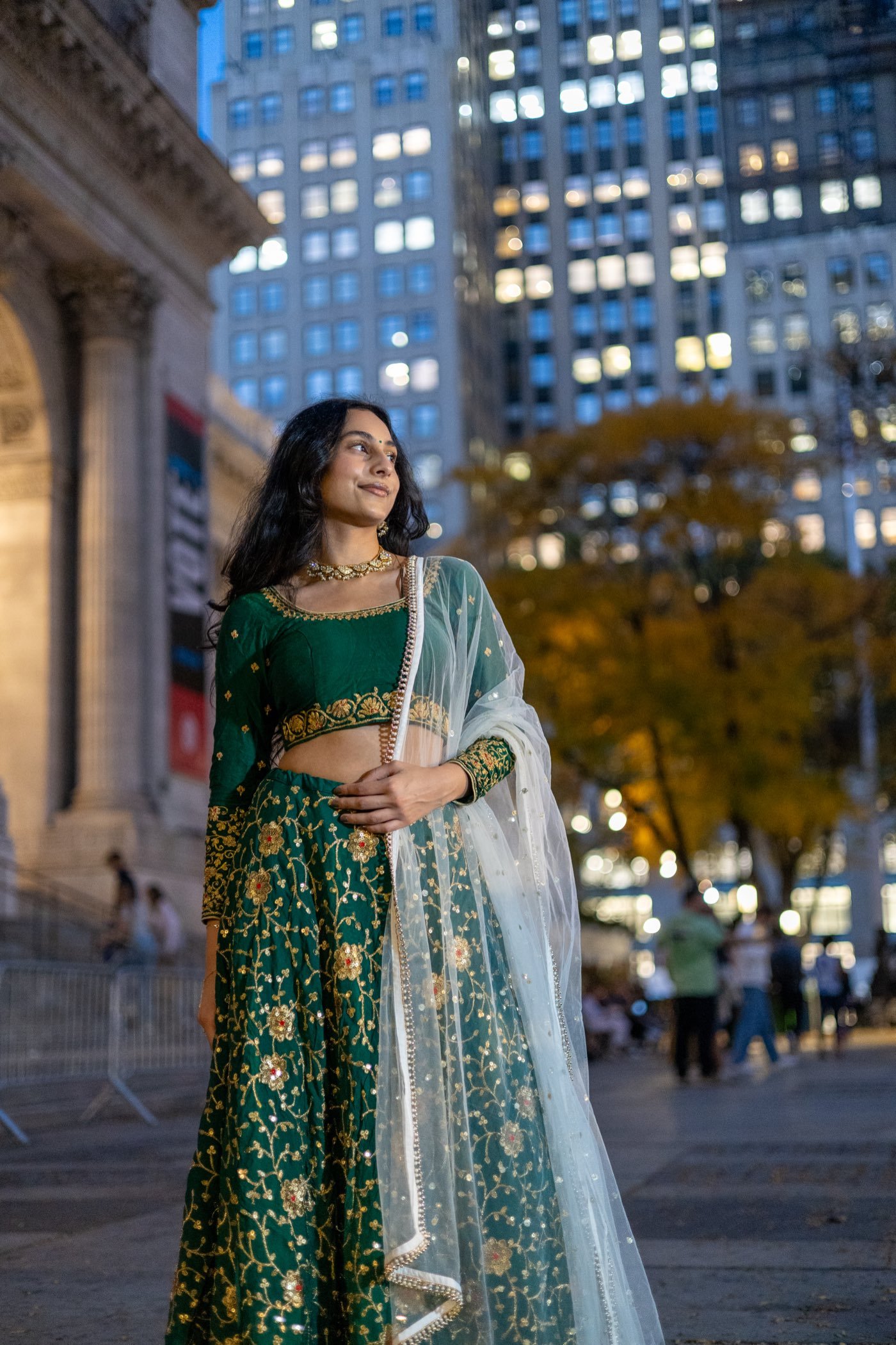 Model in an emerald and gold lehenga at dusk, city lights behind her
